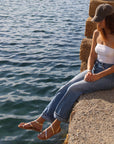 Woman wearing Theluto Lilou handcrafted gold leather sandals while sitting on stone steps by the sea