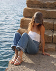 A woman wearing Theluto Lilou handcrafted silver leather sandals while sitting on stone steps by the sea.