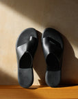 Theluto Mykonos black leather toe-loop sandals with a minimalist slip-on design on a sunlit textured wall