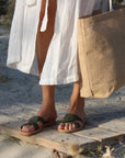 Theluto Prune olive green leather slip-on sandals worn with a white linen dress and a beige jute tote bag on sand.