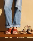 Theluto Tom minimalist leather sandals in tan worn with jeans, alongside a metallic gold pair on a wooden bench.