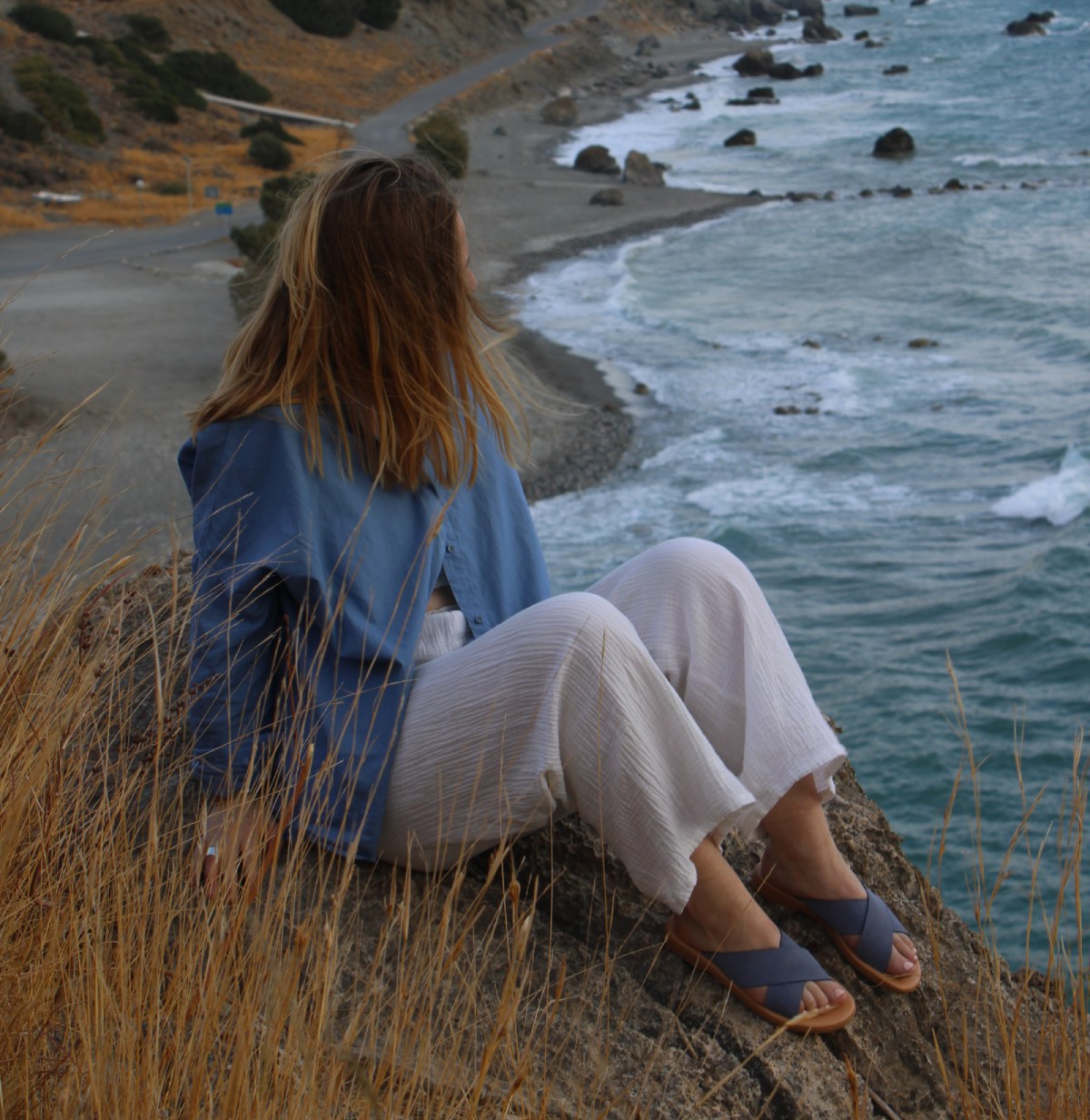 Woman wearing Theluto Gaelle leather slip-on sandals while sitting on a cliff overlooking the ocean