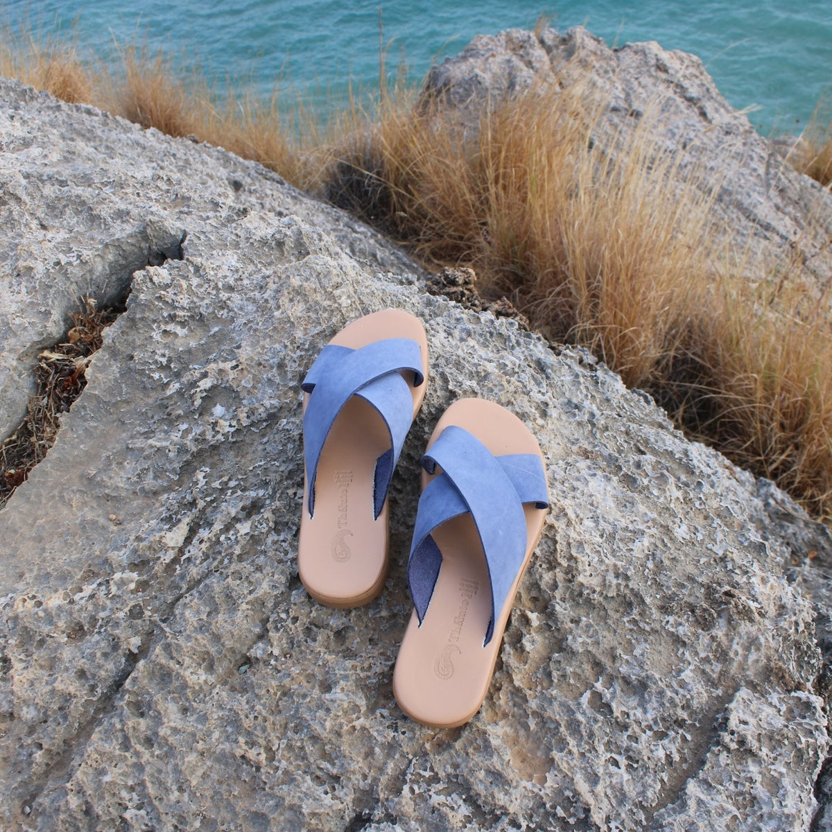 Theluto Gaelle blue leather slip-on sandals with a cushioned sole, displayed on a rocky cliff overlooking the sea.