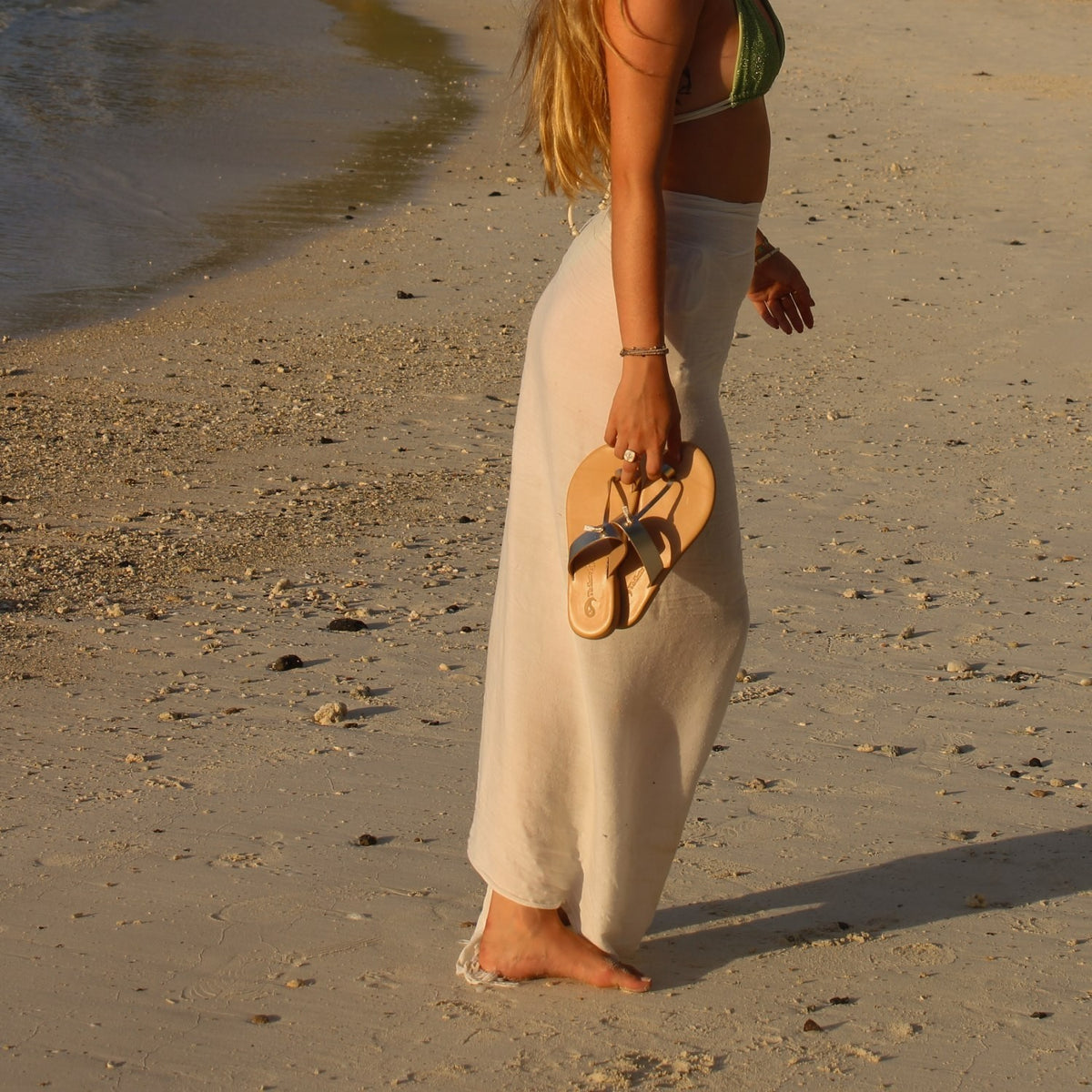 Theluto Héloïse handmade natural leather sandals held by a woman in a white sarong on a sandy beach at sunset