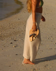 Theluto Héloïse handmade natural leather sandals held by a woman in a white sarong on a sandy beach at sunset