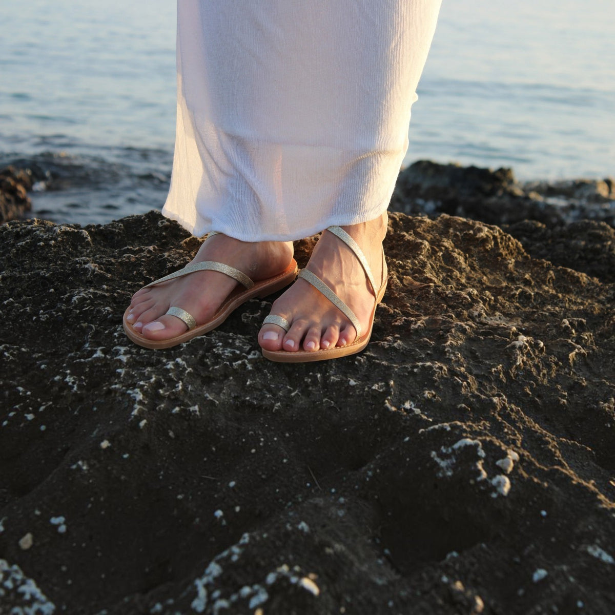 Theluto Lilou Glitter handcrafted leather sandals in gold, worn by a woman on a rocky shore.