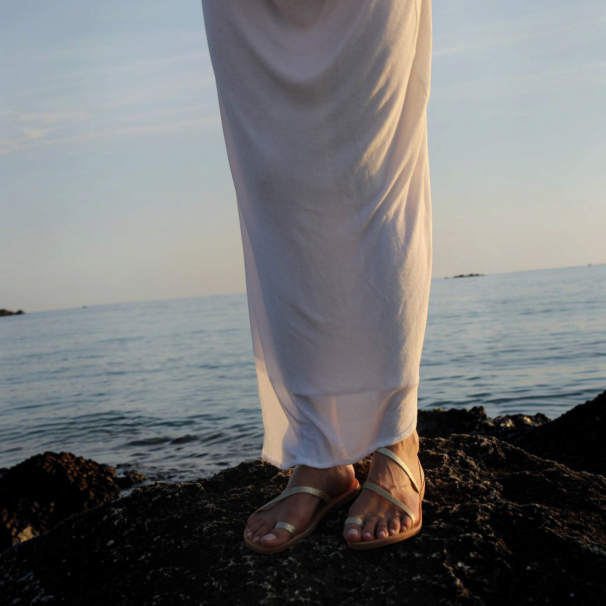 Theluto Lilou Glitter leather sandals with metallic straps worn on a rocky beach with a white dress