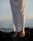 Theluto Lilou Glitter leather sandals with metallic straps worn on a rocky beach with a white dress