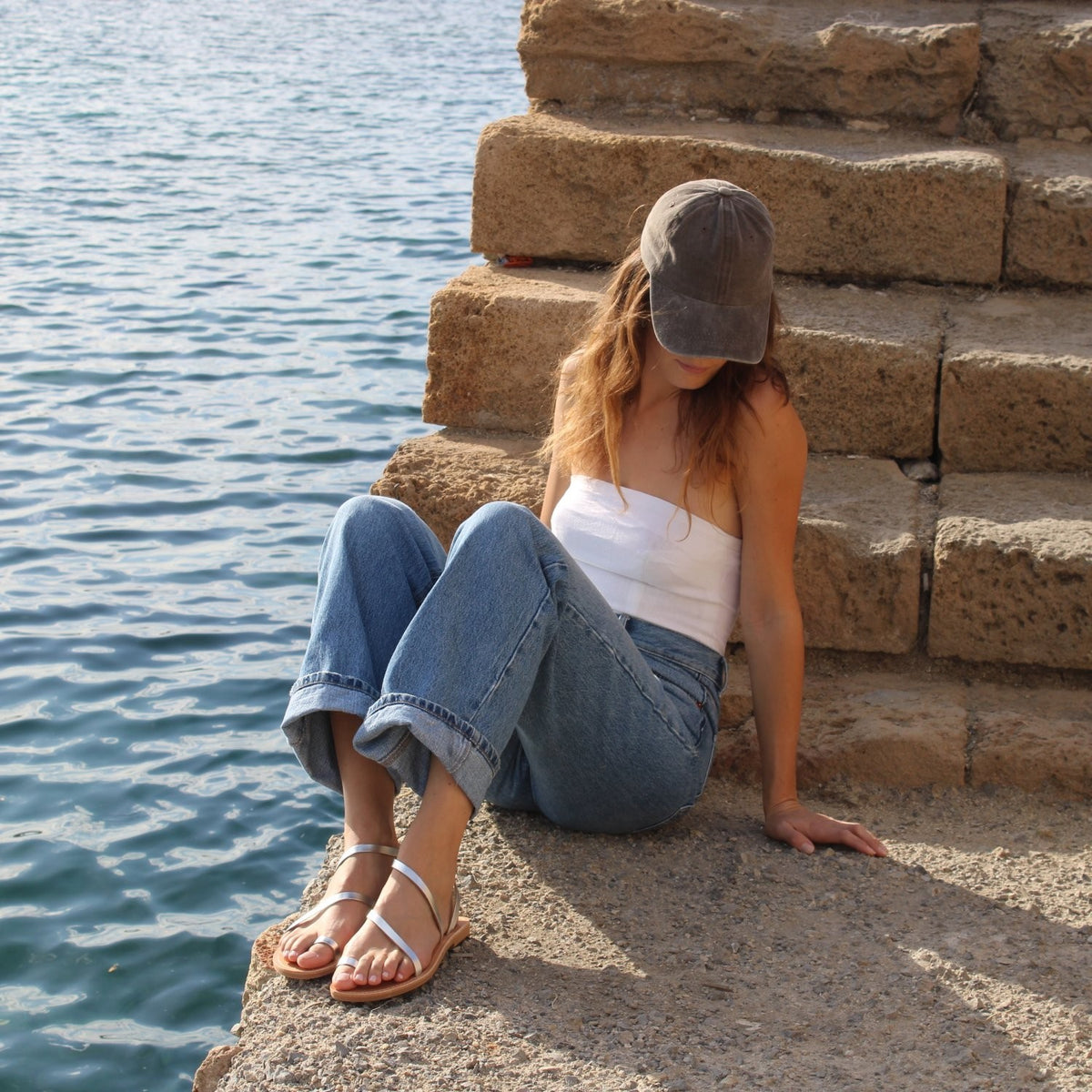 A woman wearing Theluto Lilou handcrafted silver leather sandals while sitting on stone steps by the sea.