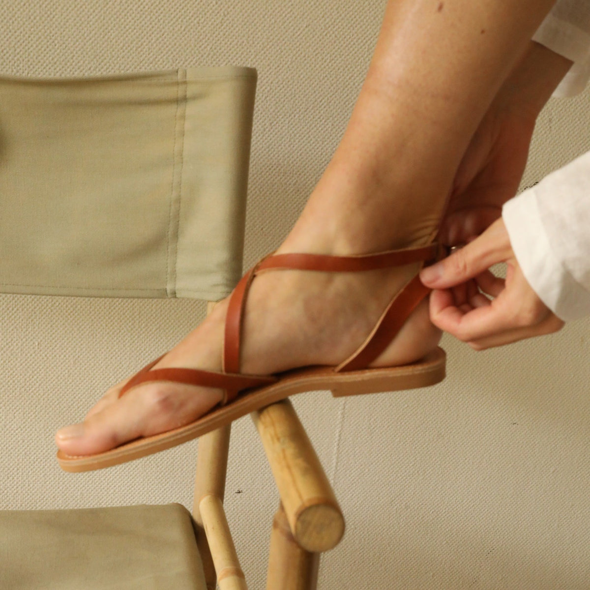 Theluto Marie tan leather thong sandals with minimalist straps being fastened on a foot while resting on a chair