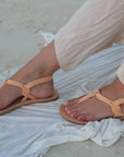 Theluto Meloe tan leather sandals with ankle ties worn on a sandy beach with light linen trousers
