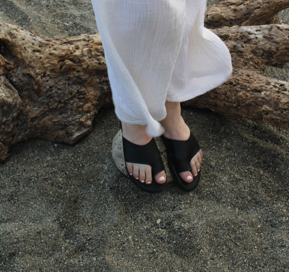 Theluto Mykonos black leather slip-on sandals worn on sandy beach with white linen trousers