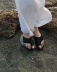 Theluto Mykonos black leather slip-on sandals worn on sandy beach with white linen trousers