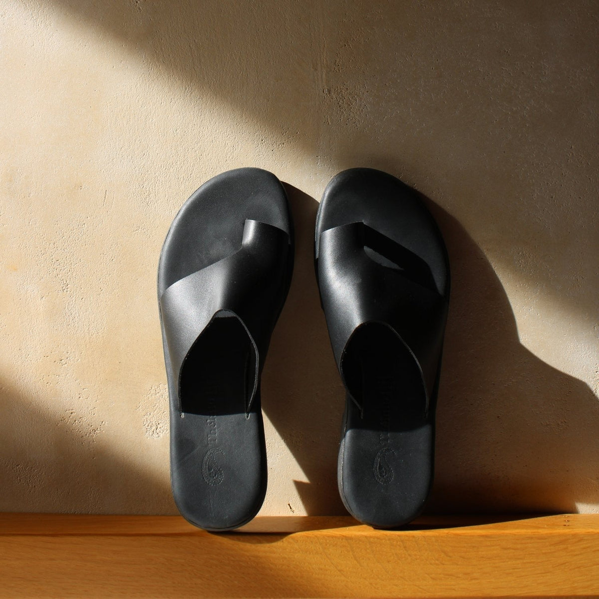 Theluto Mykonos black leather toe-loop sandals with a minimalist slip-on design on a sunlit textured wall