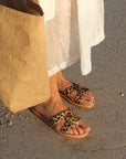 Theluto Prune Cheetah leather slip-on sandals with animal print, worn on a sandy beach with a linen dress and tote bag.