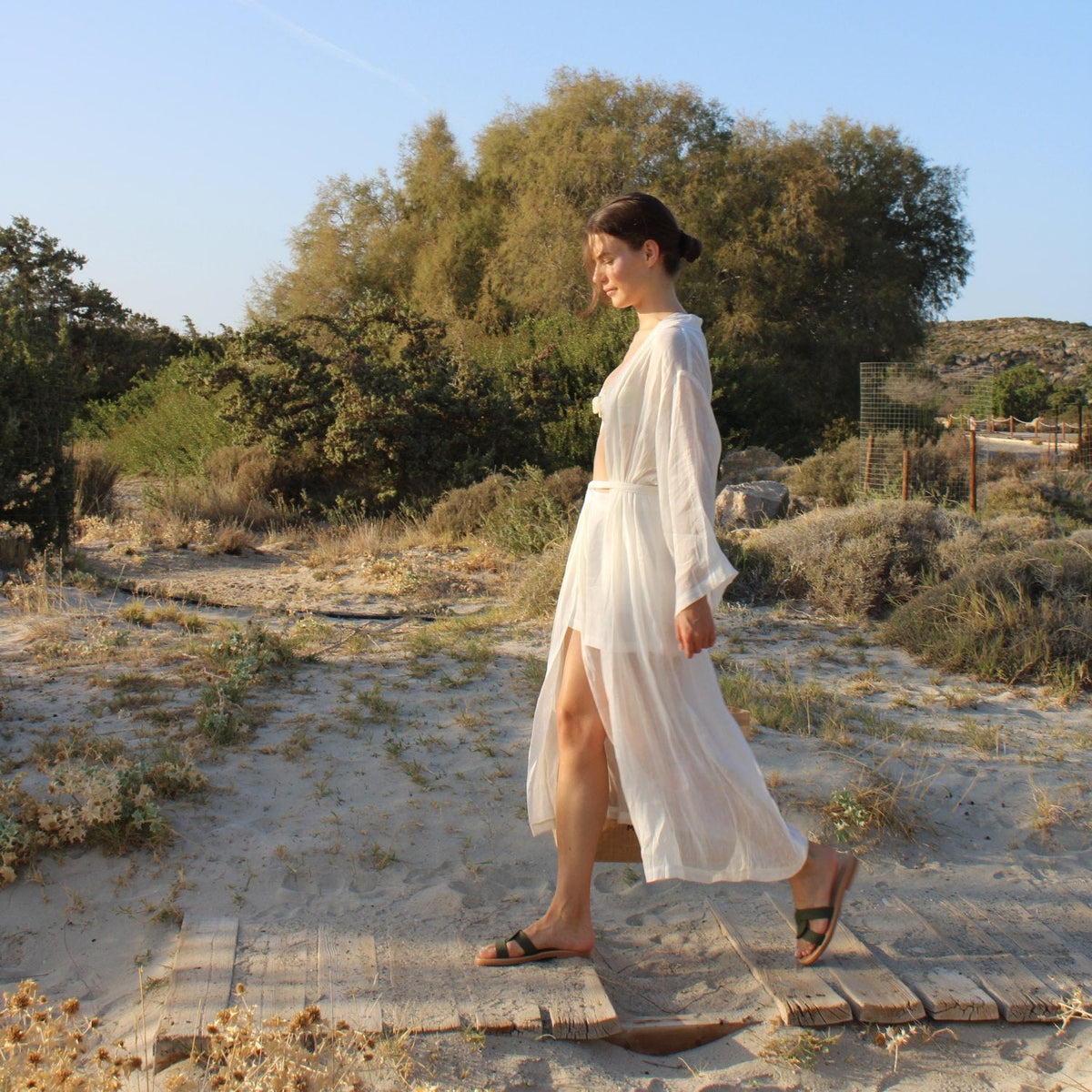 Woman wearing Theluto Prune handmade leather slip-on sandals while walking on a sandy path in a white cover-up