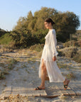 Woman wearing Theluto Prune handmade leather slip-on sandals while walking on a sandy path in a white cover-up