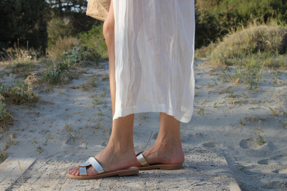 Theluto Prune handmade silver leather slip-on sandals worn by a woman in a white dress on a sandy beach path