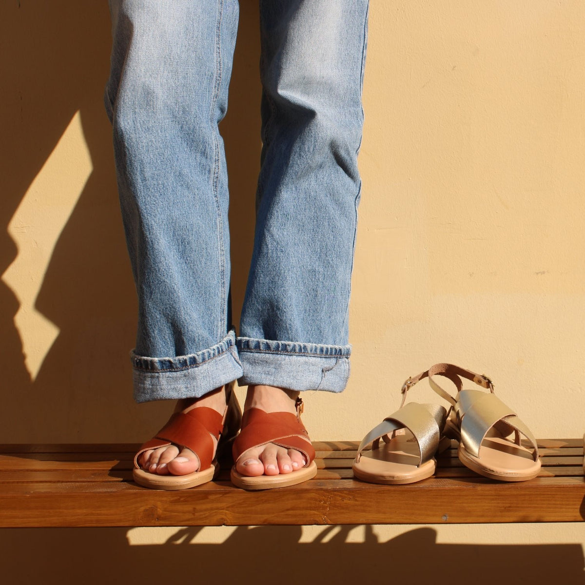 Theluto Tom minimalist leather sandals in tan worn with jeans, alongside a metallic gold pair on a wooden bench.