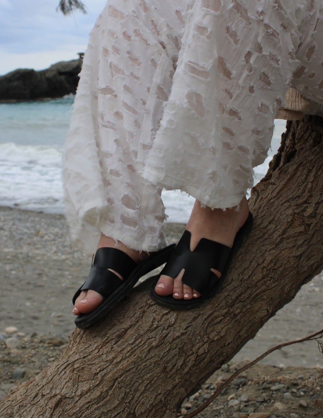 Theluto Tribeca black leather slip-on sandals worn by a woman in white trousers on a beach