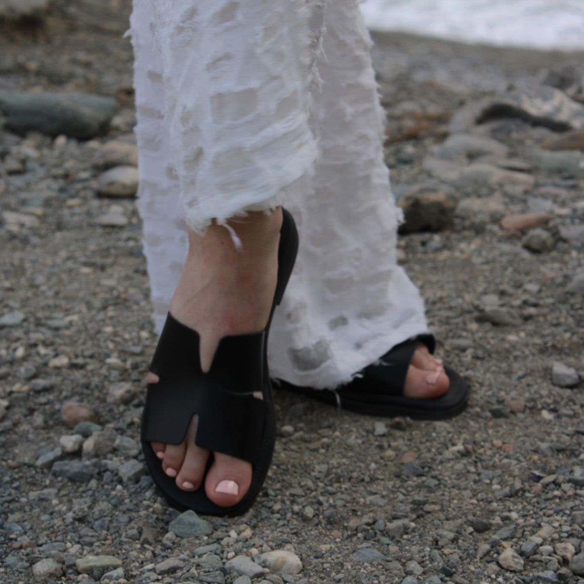 Theluto Tribeca black leather slip-on sandals worn with white distressed jeans on a rocky beach