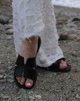 Theluto Tribeca black leather slip-on sandals worn with white distressed jeans on a rocky beach