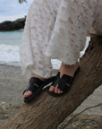 Theluto Tribeca black leather slip-on sandals worn with white textured trousers on a beach setting