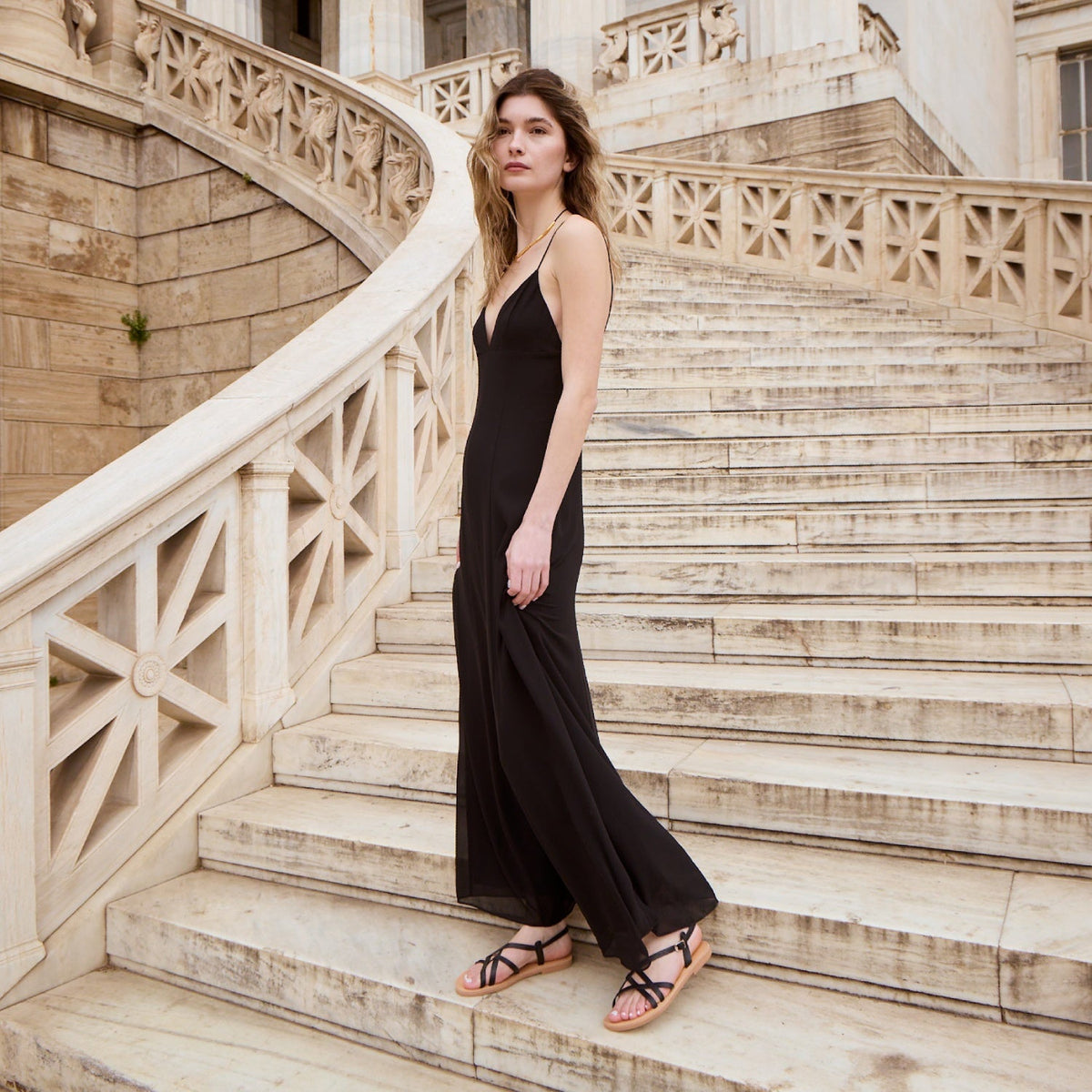 A woman wearing Theluto Valentine black leather slip-on sandals with a long black dress on stone stairs