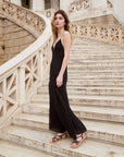 A woman wearing Theluto Valentine black leather slip-on sandals with a long black dress on stone stairs