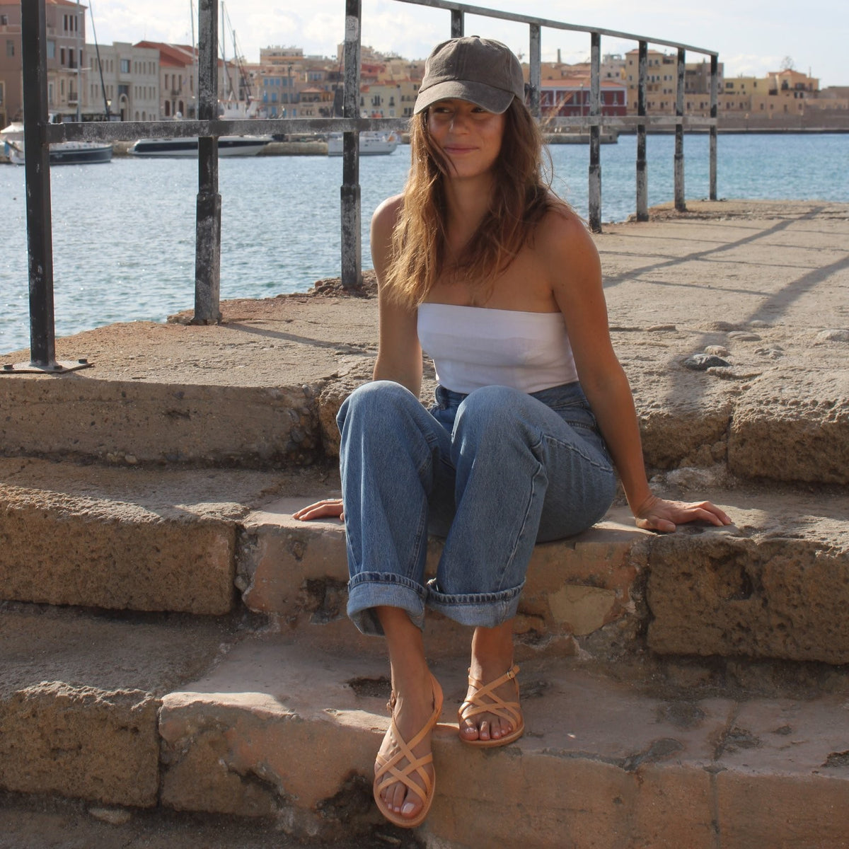 A woman wearing Theluto Valentine tan leather slip-on sandals while sitting on stone steps by a harbor.