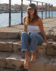 A woman wearing Theluto Valentine tan leather slip-on sandals while sitting on stone steps by a harbor.
